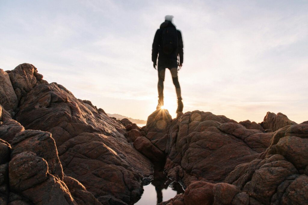 Full body unrecognizable tourist in black jacket with backpack standing on rocky shore and admiring sunrise